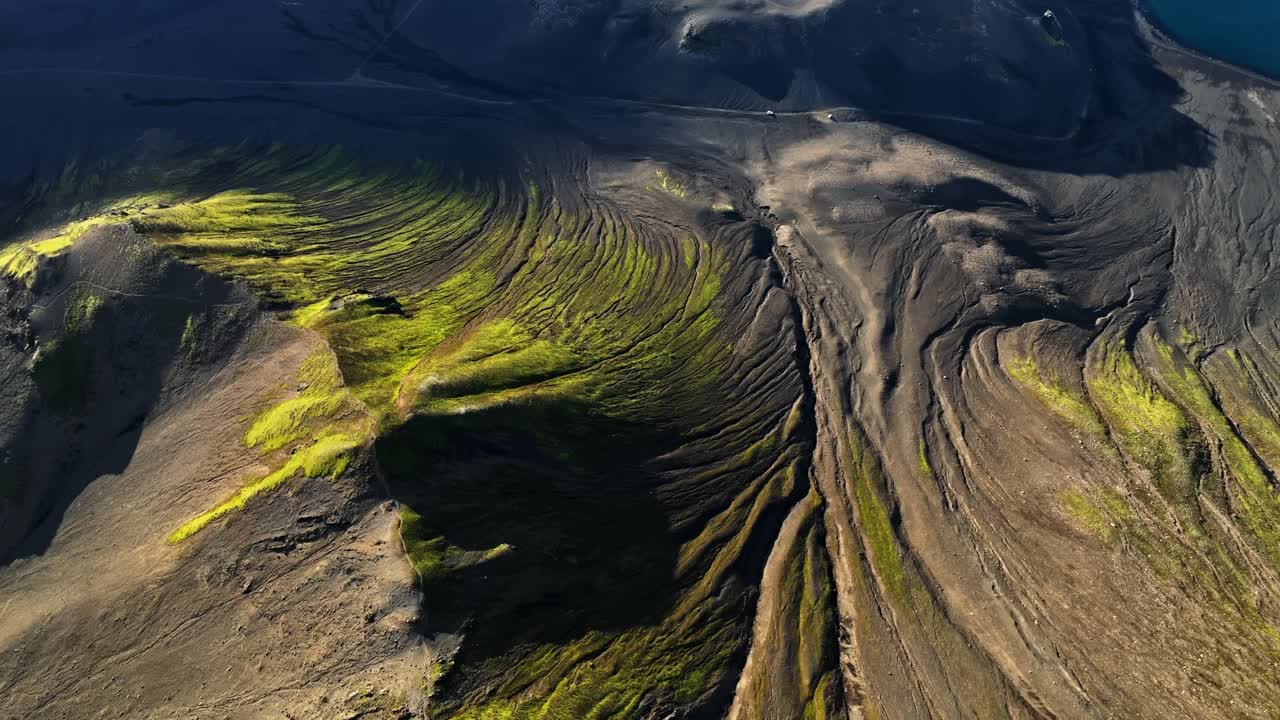 Aerial view of sweeping glacial river beds carved into volcanic slopes, where dark sediment channels and bright moss streaks reveal centuries of meltwater shaping Iceland’s rugged highlands