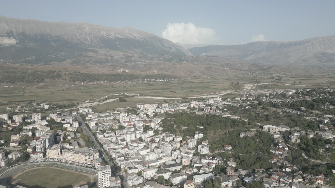 Ripresa drone sopra Gjirokastra, Albania, in una giornata di sole con foschia, la città di case bianche, montagne e nuvole sullo sfondo. LOG