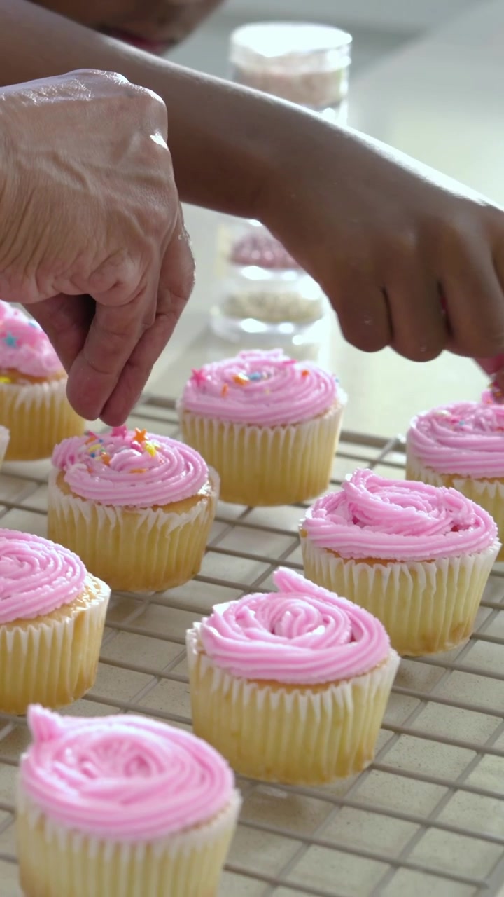 Vídeo vertical: Avó orientando no balcão, menina de camiseta verde colocando granulado em cupcakes.