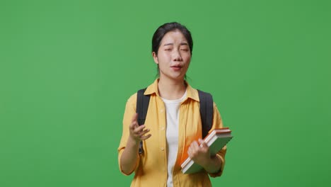 녹색 스크린 배경 스튜디오에 서 있는 동안 카메라를 보고 누군가에게 소리치는 배과 몇 권의 책을 가진 아시아 여성 학생