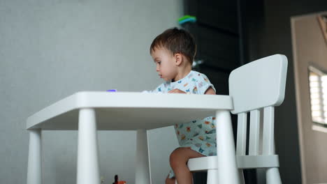 focused baby boy sits at white desk in his room. kid plays with his toys cheering and clapping hands.
