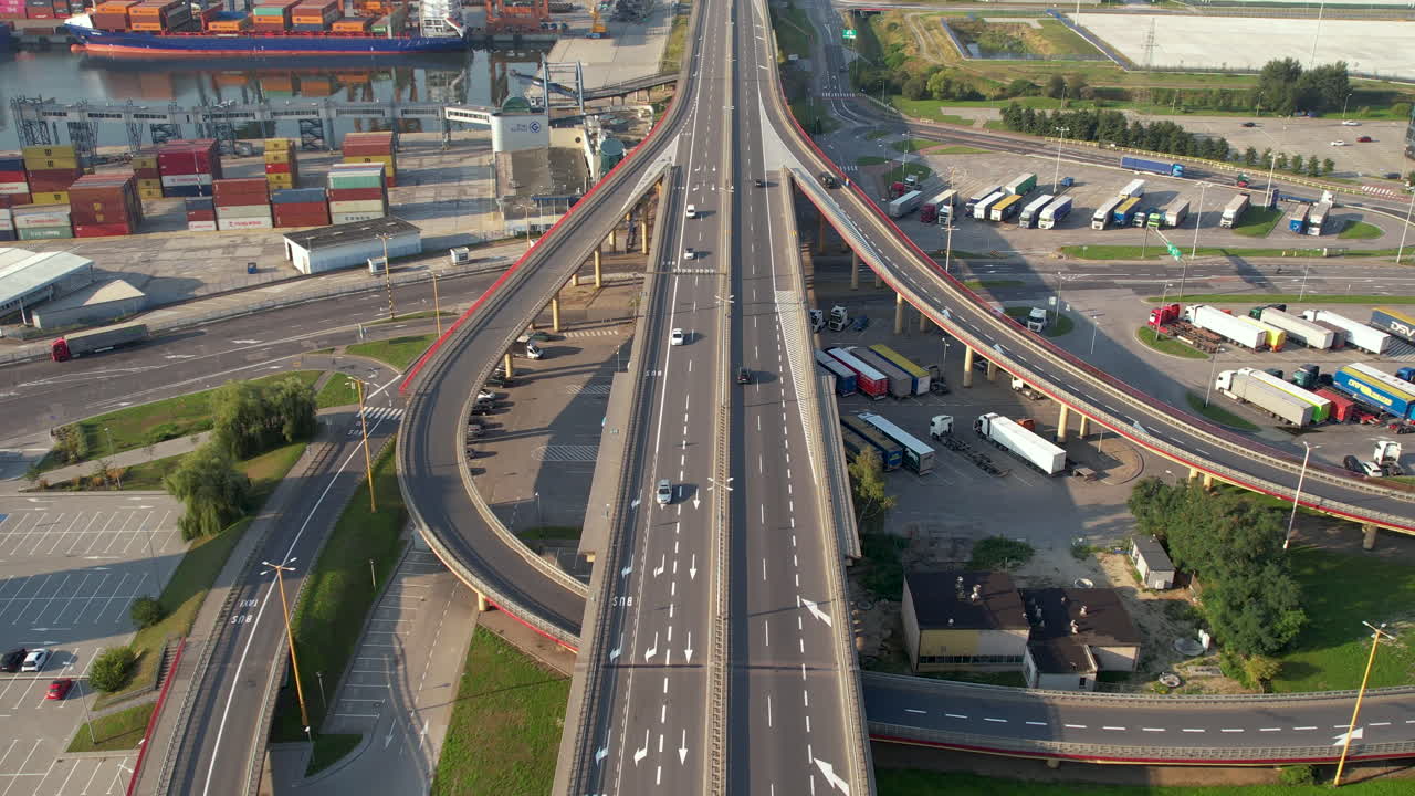 Pengambilan gambar tilt-up udara mobil-mobil yang melaju di jalan raya dekat Pelabuhan Gdynia dengan kapal kontainer yang sedang berlabuh, Polandia.