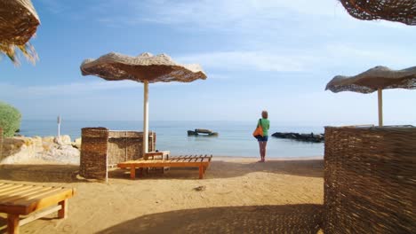 Steadicam-Shot-Woman-Standing-On-The-Beach-Near-The-Parasols-And-The-Palm-Trees-Looking-At-The-Sea