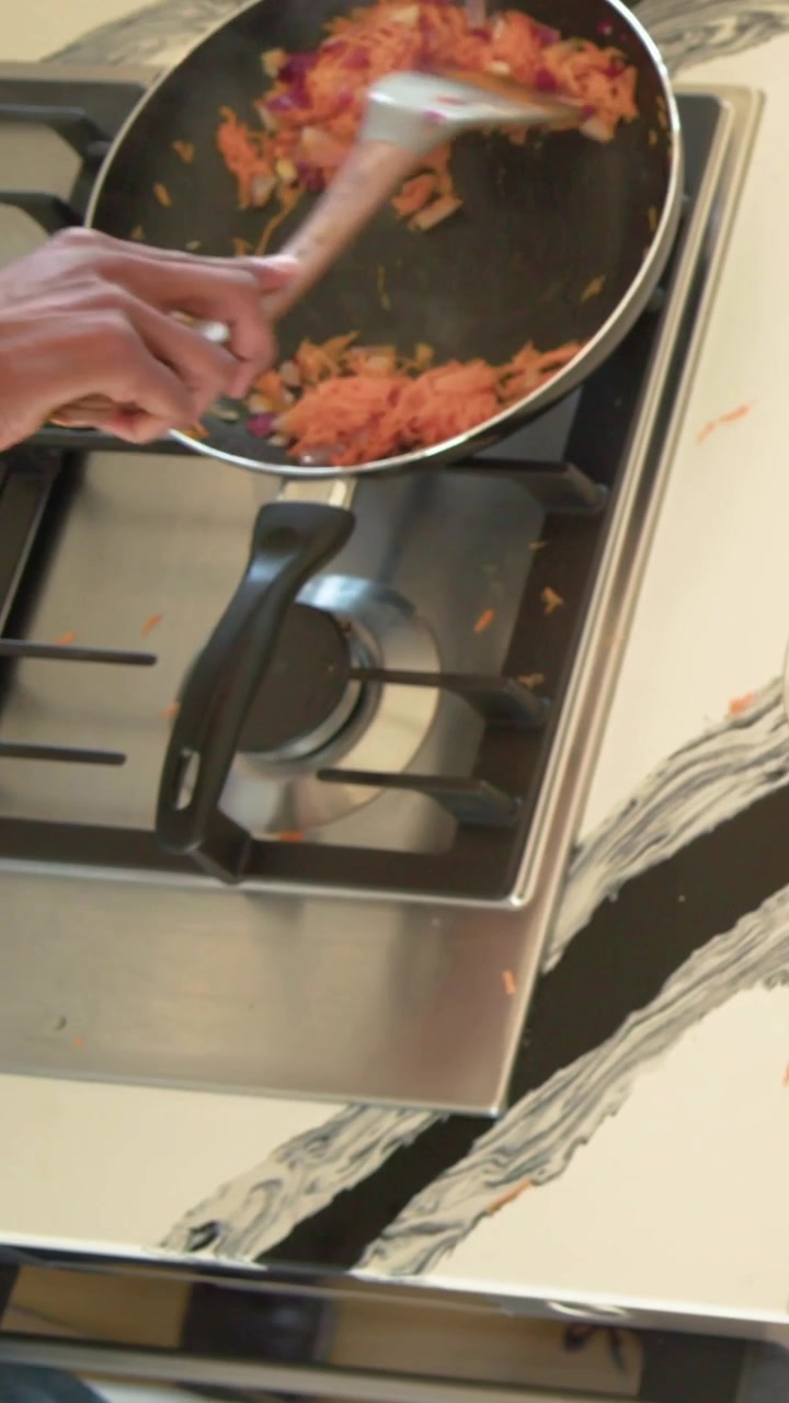 Vídeo vertical: Cozinheiro(a) doméstico(a) com blusa verde pegando cenouras raladas e mexendo no fogão com uma espátula.