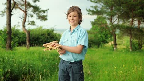 close-up view of happy little boy playing with wooden airplane toy and smiling at camera in the park on a cloudy day