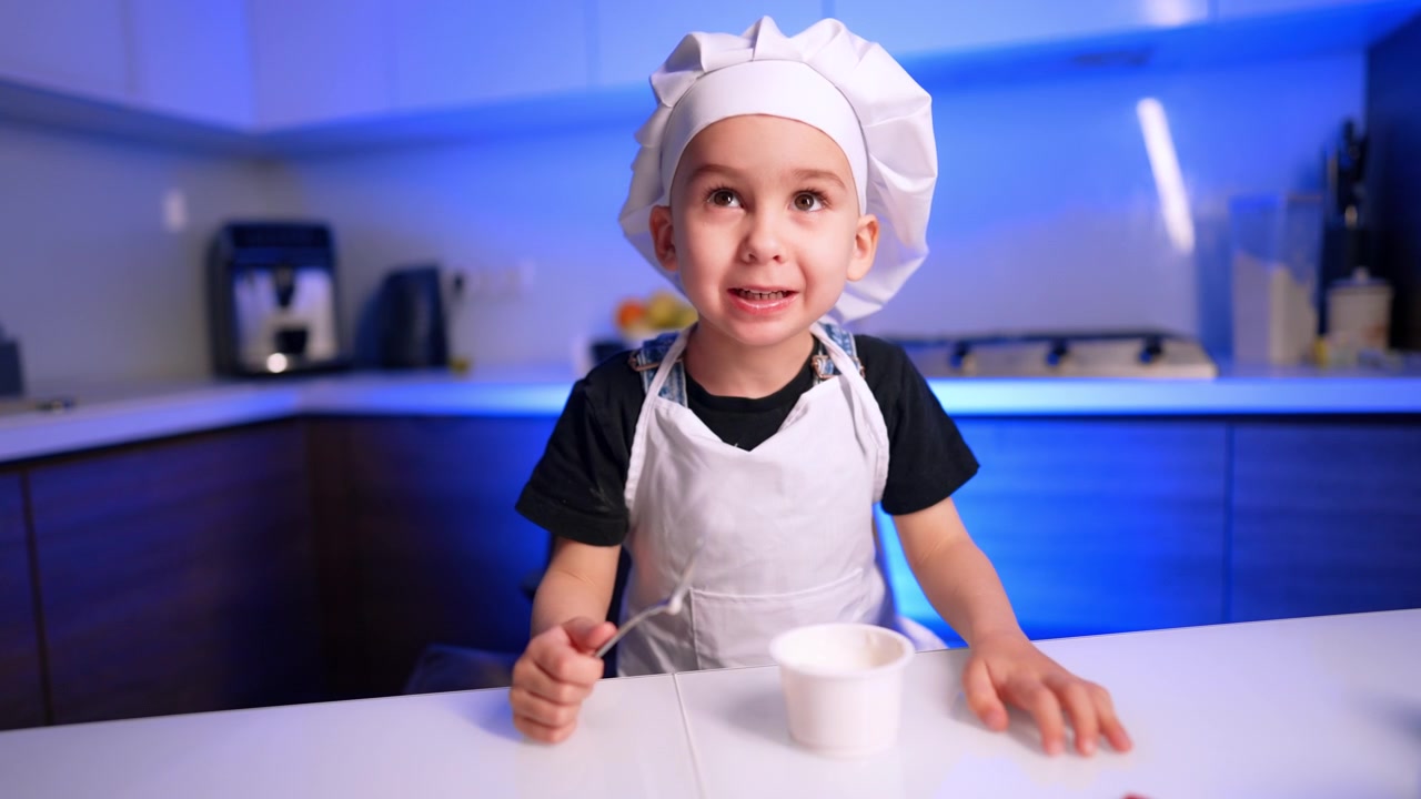 Bambino che assaggia il cibo da un contenitore di plastica bianco. Il bambino, con cappello da cuoco e grembiule, tiene un cucchiaio sopra una tazza in cucina