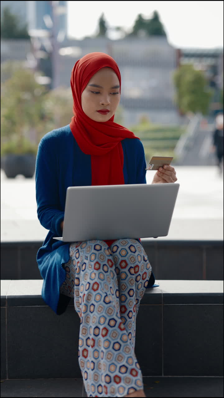 Young woman in hijab using a laptop and credit card for online shopping or payment outdoors