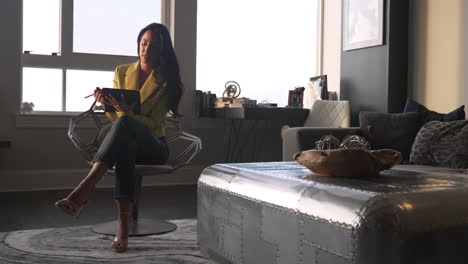 Young-urban-businesswoman-contemplating-and-writing-on-her-digital-tablet-in-her-penthouse-living-room-home