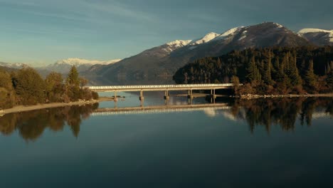 aerial shot orbiting around a bridge in the middle of a scenic lake with mountains on background near bariloche city, argentina