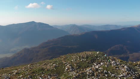 three-hikers-reach-top-of-high-mountain,-drone-flies-over-edge