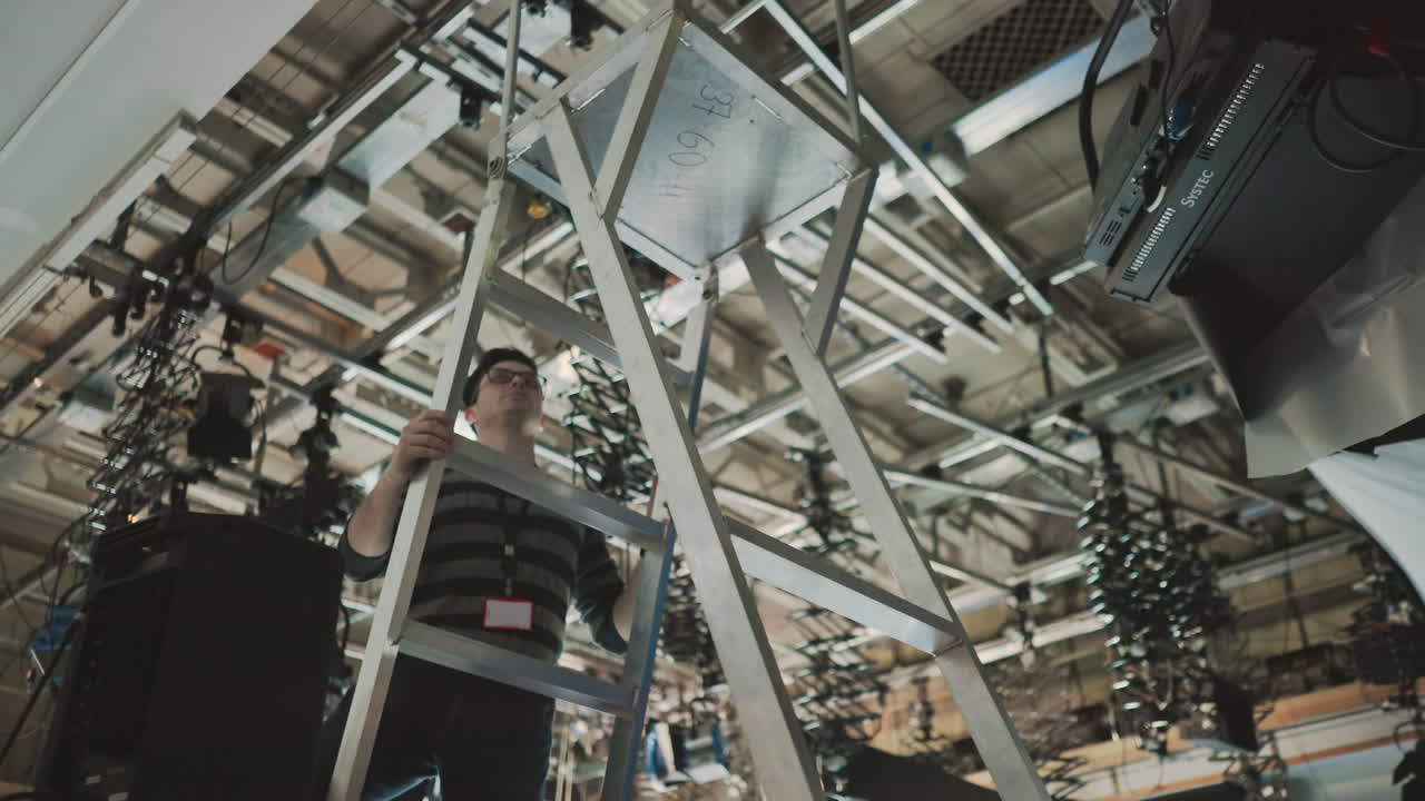 Worker climbing metal ladder in studio environment, focusing on ceiling mounted lights and camera equipment, preparing for shooting or lighting adjustments