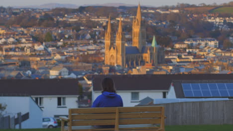 person sits on bench overlooking truro, uk cathedral at sunset during lockdown, zoom in