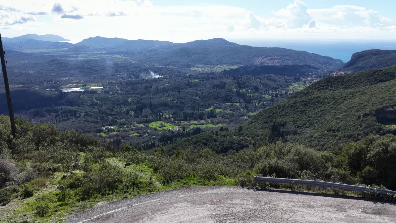 Vista aérea de drone sobre uma estrada de montanha, revelando um vale verdejante, uma vila tradicional e um mar distante em Corfu, Grécia. Imagens de tirar o fôlego da paisagem natural da ilha mediterrânea.