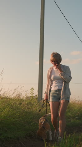 woman in jean shorts walking on phone call during early morning through grassy field, holding leash in one hand, with background featuring greenery, electric pole, and distant rural building