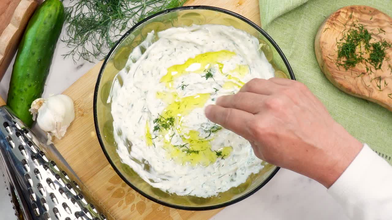 Making Tzatziki Greek or Mediterranean sauce or dip for a party. Dip or sauce in a green bowl on a wooden cutting board with dill, green napkin and a grater. Adding dill to the top of Tzatziki sauce.