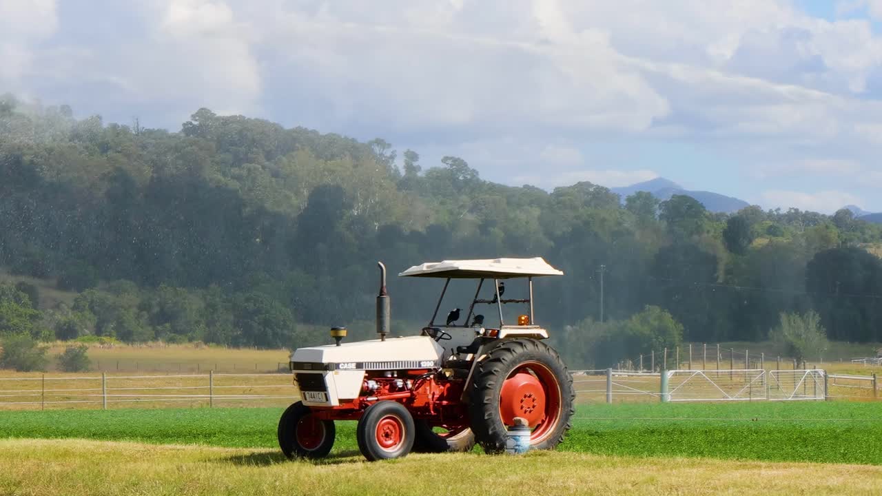 A red tractor sprays water across a vibrant green field under a cloudy sky.