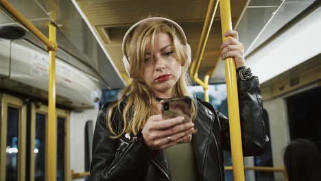 young tired girl hipster in headphones is listening to music. riding tram, using smartphone, standing and holding onto the handrail. slow motion