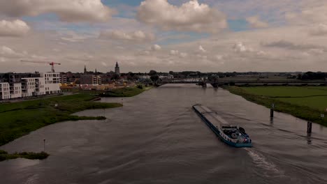 Großes-Frachtschiff-Auf-Der-Ijssel,-Vorbei-An-überschwemmungsgebieten-Und-Zeitgenössischen-Modernen-Konstruktionen-Am-Flussbett,-Das-In-Die-Historische-Hansestadt-Zutphen,-Niederlande,-Einfährt