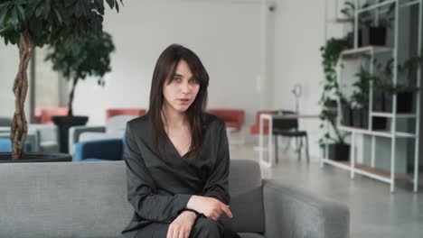 young,-beautiful-woman-in-a-suit-scolds-while-looking-at-the-camera-with-a-sorrowful-expression-on-her-face-and-gesticulates,-sitting-on-the-couch-in-the-business-center-lobby