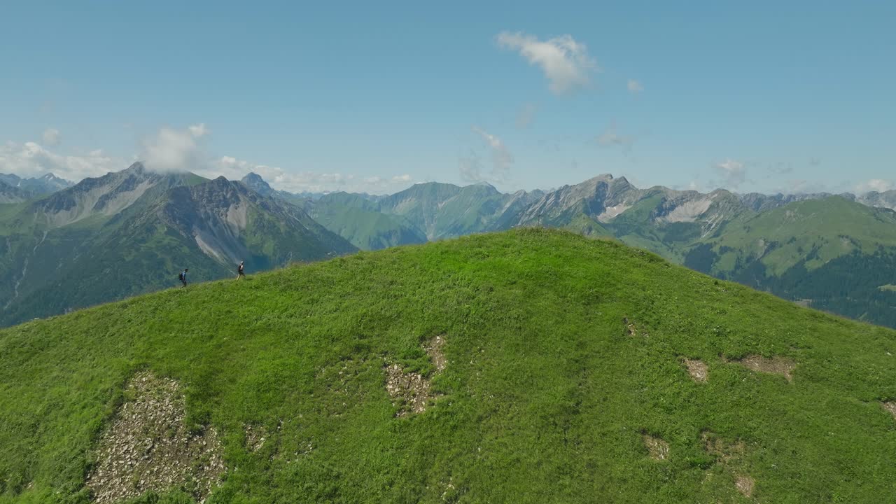 Two hikers trek a grassy hilltop with a vast mountain range in the background under a clear sky