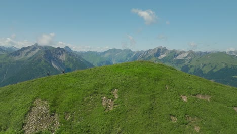 two hikers trek a grassy hilltop with a vast mountain range in the background under a clear sky