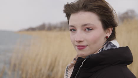 Close-Up-View-Of-Teenage-Girl-With-Short-Hair-Wearing-Scarf-And-Coat-Looking-At-Camera-And-Smiling-On-A-Windy-Day