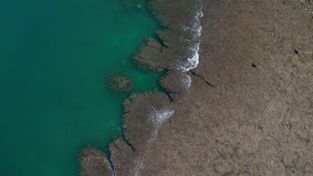 A captivating drone shot captures the surface of the shore, where the ocean’s waves gently crash, leaving behind delicate white foam that clings to the golden sands.