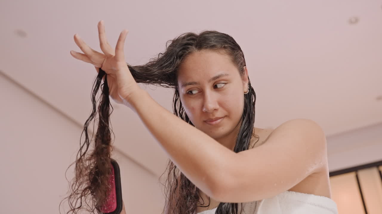 Preparándose para la noche con el cabello recién peinado. Mujer envuelta en una toalla arregla cuidadosamente su largo cabello húmedo. Mujer en toalla peina meticulosamente su cabello largo y húmedo antes de salir por la noche.