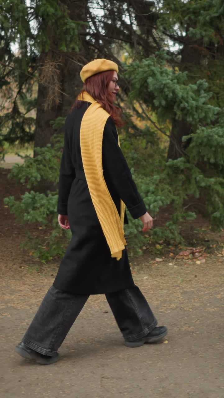 Side view of an elegant lady with long wavy red hair, wearing a black coat, yellow beret, and matching scarf, joyfully walking through a lush green park adorned with colorful autumn foliage