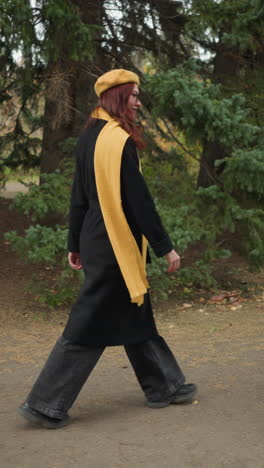 vista lateral de una dama elegante con el cabello rojo largo y ondulado, con un abrigo negro, boina amarilla y bufanda a juego, caminando alegremente a través de un exuberante parque verde adornado con coloridos follajes de otoño