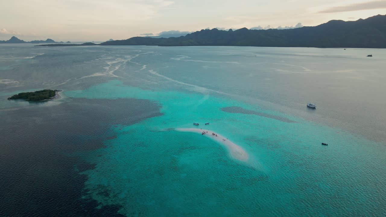 A narrow white sandbar stretches across crystal-clear turquoise waters in Komodo Island, surrounded by deep blue sea. Boats anchor nearby, with island hills silhouetted in the distance.