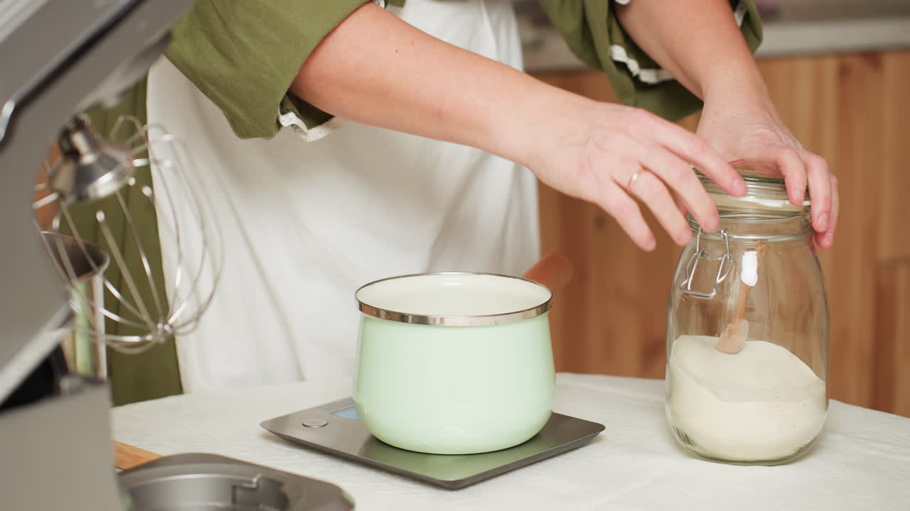 Baker in green top and white apron opens flour jar, scoops flour with wooden spoon, and gently pours it into mint green pot placed on digital kitchen scale during precise baking preparation