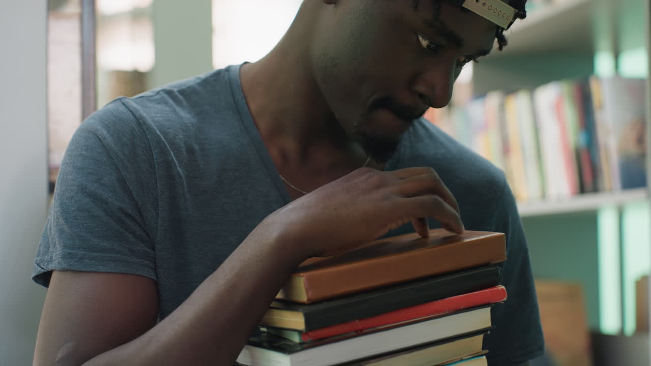 Close up of young man in casual shirt carrying stacked books in library, gently tapping cover of top book while appearing deep in thought, surrounded by blurred shelves filled with colorful books