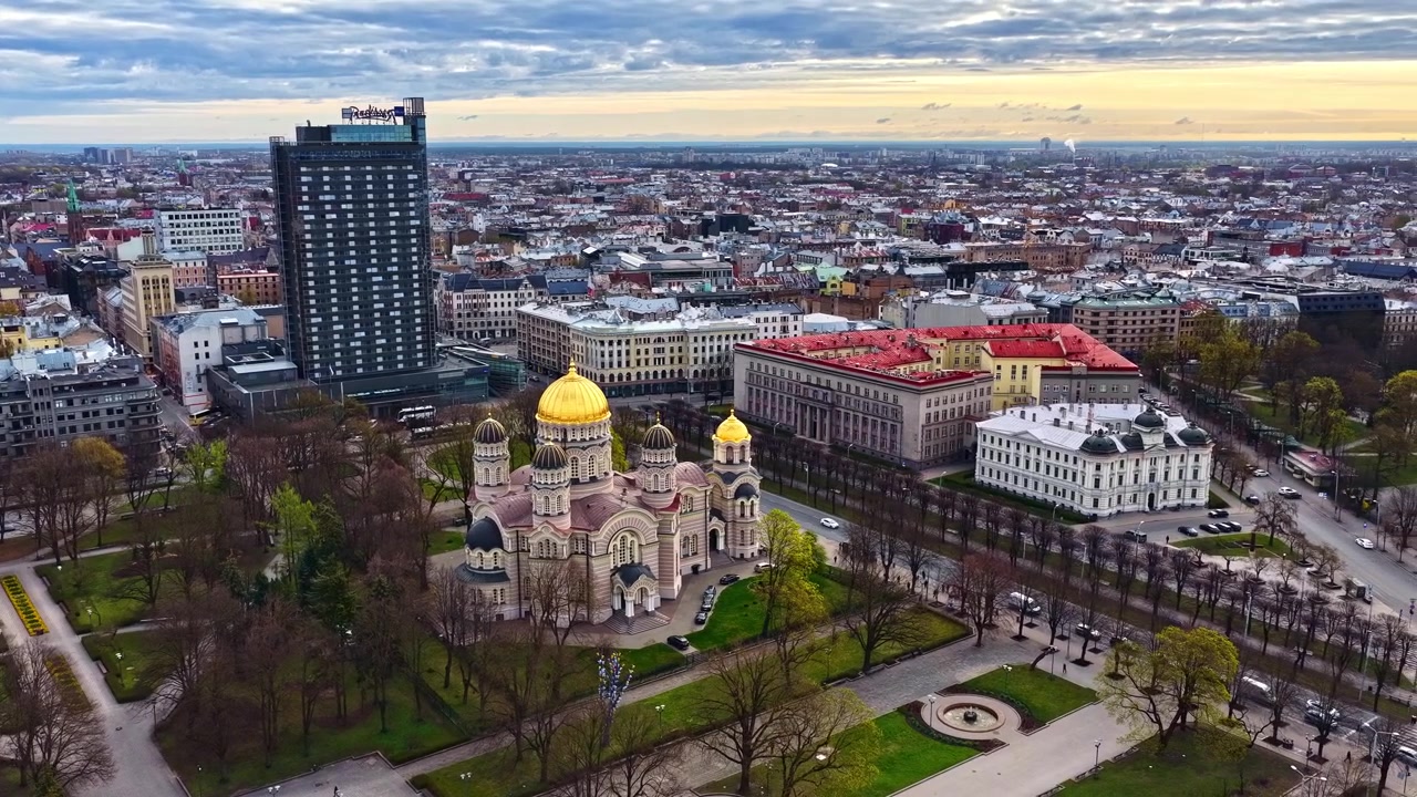 Vista aérea sobrevoa o centro de Riga, mostrando parques, edifícios históricos e modernos ao anoitecer.