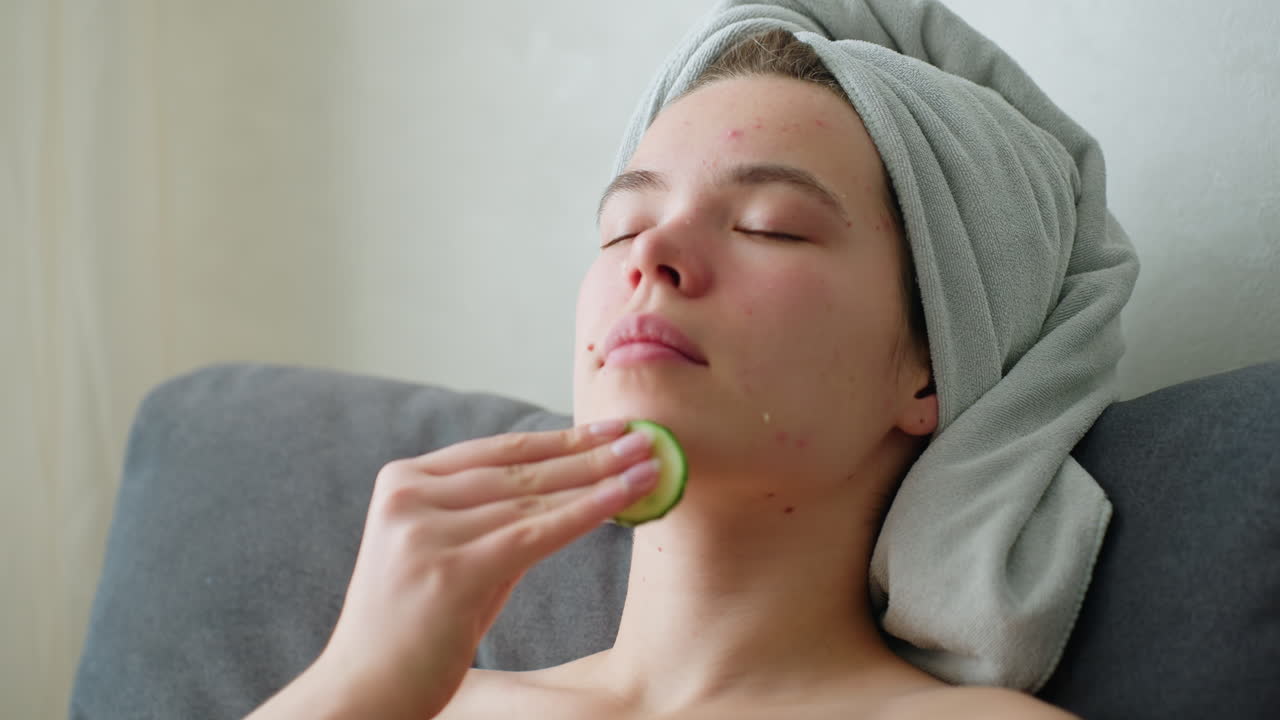 Close-up of woman with towel around head gently rubbing cucumber slice on her cheek during skincare routine while relaxing in bright indoor setting, promoting freshness, calmness