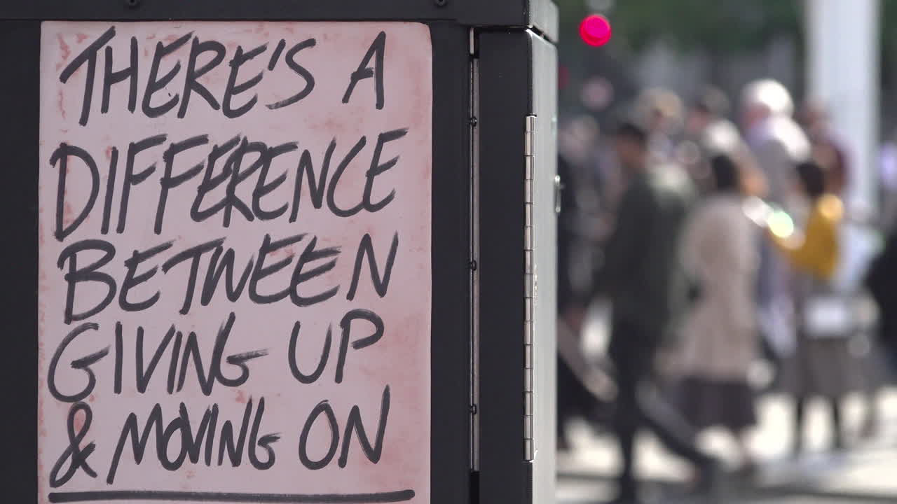 UK October 2018 - Out of focus people on a street walk past a homemade fly poster sign on an electrical junction box that says, “There’s a difference between giving up and moving on”