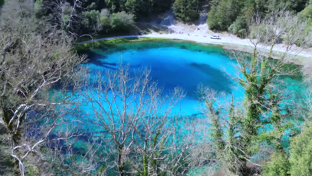Cinematic 6-second drone shot of the crystal clear "Blue Eye" springs of Louros. High-angle view of the vibrant turquoise circular lake nestled in a lush green forest. Perfect for nature intros