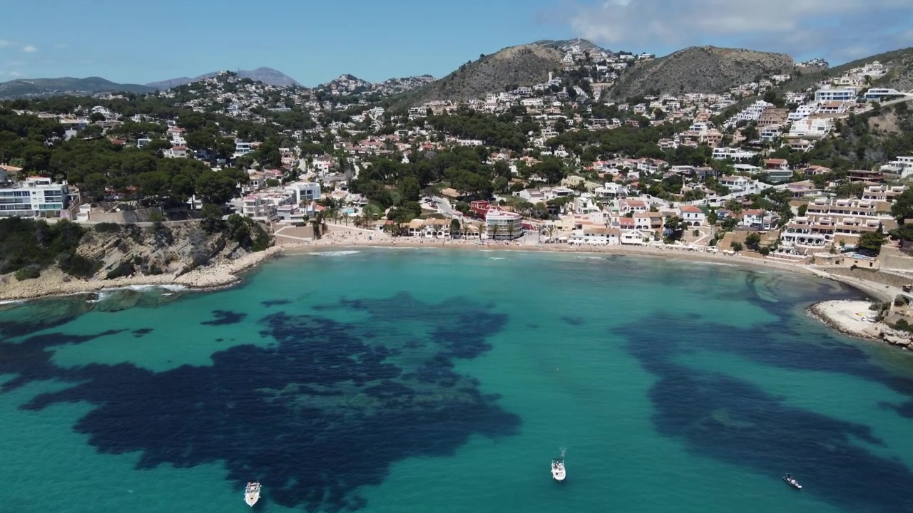 Splendida vista aerea con drone di barche ancorate che galleggiano nelle acque cristalline della spiaggia di Cala El Portet, con una maestosa catena montuosa sullo sfondo. Moraira, Costa Blanca, Spagna.