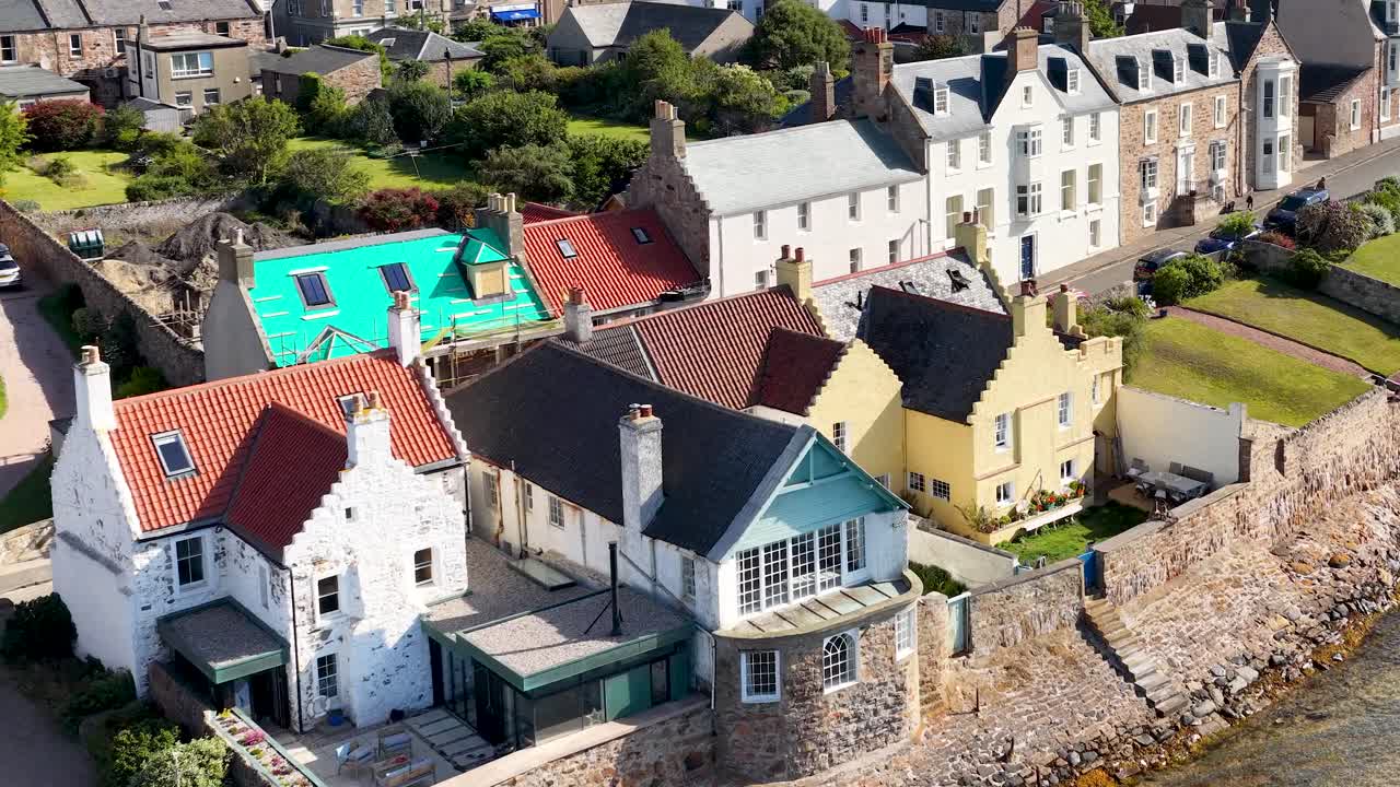 Drone glides above seaside village rooftops, historic architecture, gardens, and stone seawall in daylight