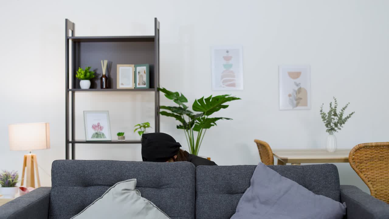 Ladrona asiática con gorro negro busca en la sala de estar mientras se esconde detrás de los muebles