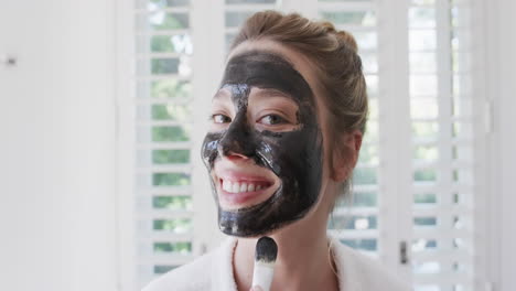 Portrait-of-happy-biracial-woman-applying-beauty-face-mask-in-bathroom,-slow-motion