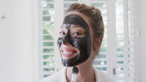 retrato de una feliz mujer biracial aplicando una máscara facial de belleza en el baño, cámara lenta