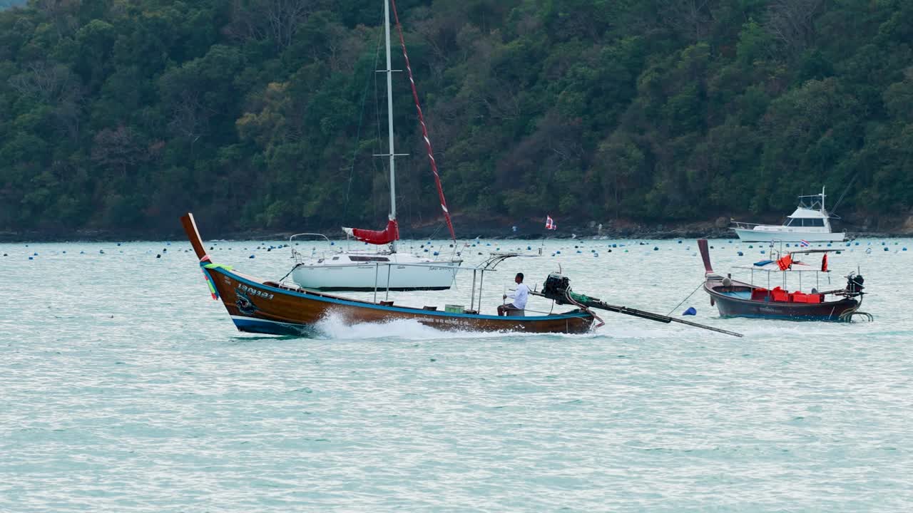 A longtail boat swiftly moves across a bay in Phuket, Thailand, under overcast skies, surrounded by anchored boats
