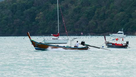 a longtail boat swiftly moves across a bay in phuket, thailand, under overcast skies, surrounded by anchored boats