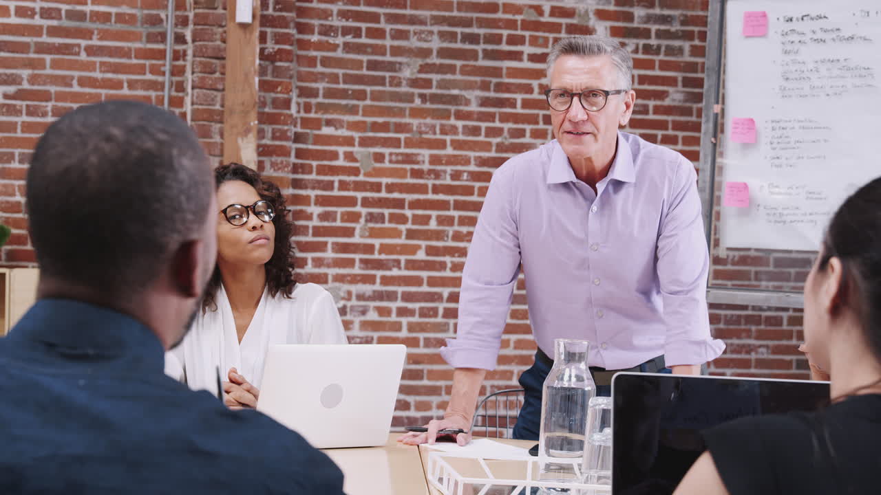 성숙 한 사업가 가 테이블 주위에 앉아 있는 동료 들 의 사무실 모임 을 이끌고 있다