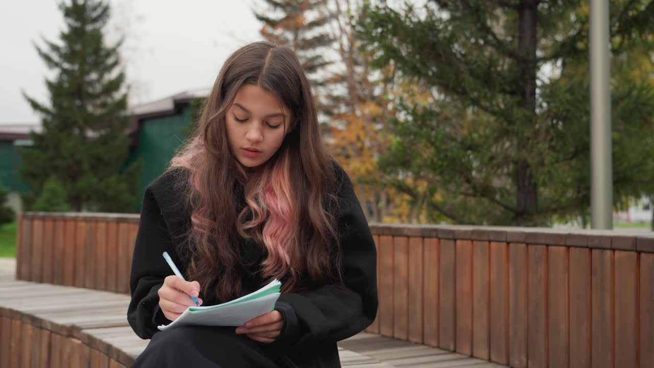 Studente adolescente che studia all'aperto su una panchina, ambientazione autunnale in un parco con ringhiera in legno e abeti, scrive su un quaderno ad anelli mentre si ferma a pensare e mordicchia la penna, indossa un maglione scuro e un cappotto informale
