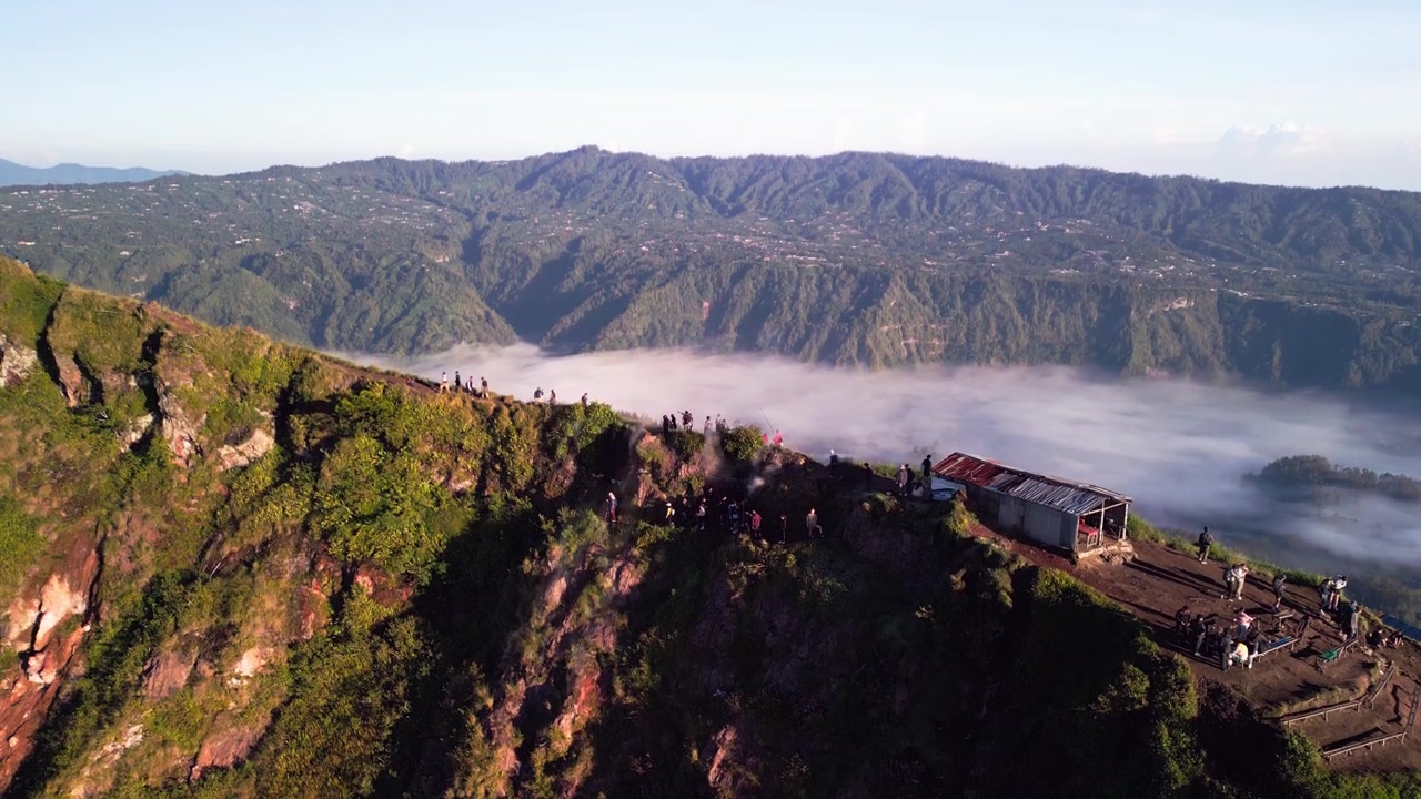 Sentiero sulla cresta del vulcano di Bali con turisti che camminano lungo il bordo del cratere, fumo vulcanico che sale dal terreno in pieno giorno con terreno accidentato ad alta quota, riprese aeree ascendenti con drone.