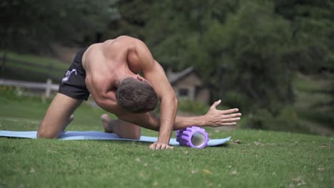 Young-caucasian-fit-attractive-male-exersicing-outdoors-Foam-roller-back-exercise-with-green-vegetation-background-slow-motion-60-fps