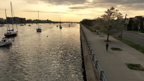 an aerial shot over sheepshead bay during a golden sunrise with boats anchored and a jogger running on the paved walkway along the water