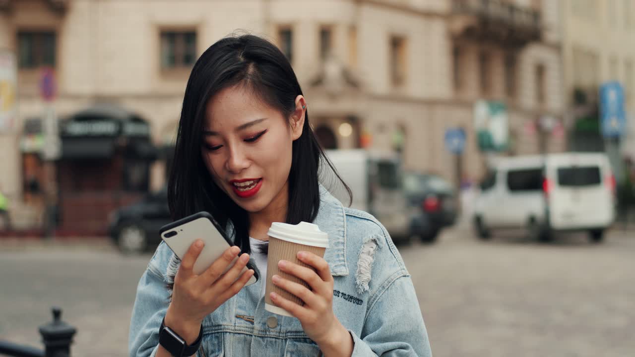 Cheerful Young Stylish Girl Chatting And Texting Messages On The Smartphone While Standing In The Center City With A Cofee To Go In Hand And Yellow Bus Going Behind Her